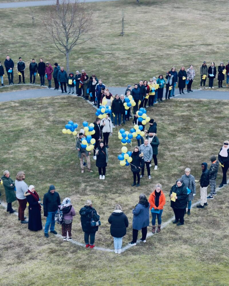 Många människor som bildar ett hjärta, där några står i mitten med gula och blå ballonger