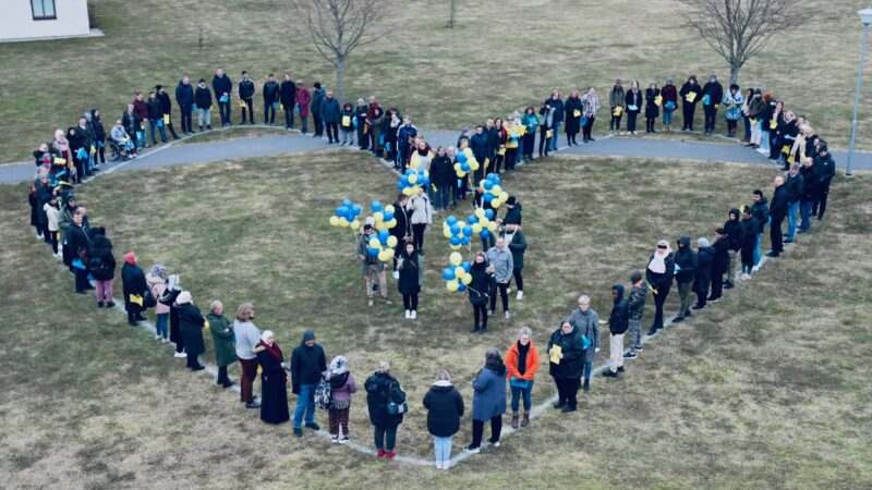 Många människor som bildar ett hjärta, där några står i mitten med gula och blå ballonger